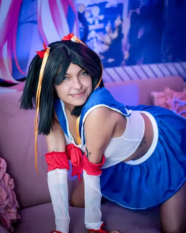 SakuraTao wears a blue and white schoolgirl outfit with red bows and long black hair while posing on a purple couch.