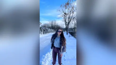 Smiling woman in a grey sweater and brown trousers enjoying a sunny winter day in a snowy landscape.