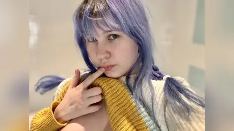 Young woman with lavender hair in a cozy yellow sweater posing in a bright, minimalist indoor setting.