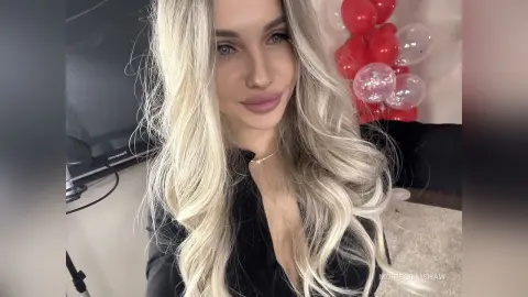 Smiling blonde woman in a black top posing in front of red balloons for a festive celebration.