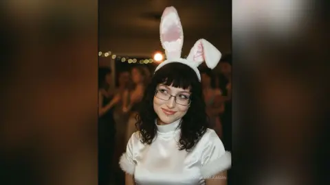 Smiling woman with glasses wearing white bunny ears at a dimly lit indoor party.