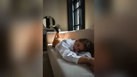 Relaxed woman in a white button-down shirt lounging on a beige sofa in a sunlit, moody living room.