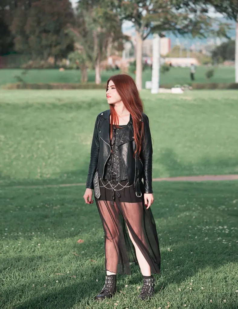 cherrryross wears a black leather jacket and corset top while standing outdoors in front of a green grass background.