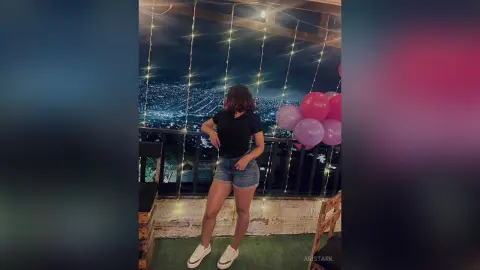 Young woman in black t-shirt and denim shorts standing on a balcony overlooking a glowing city at night.