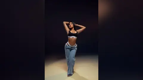 Confident woman in a black sports bra and baggy blue jeans posing against a dark studio background.