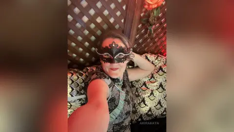 Smiling woman in a patterned dress and black masquerade mask relaxing on a patterned sofa indoors.