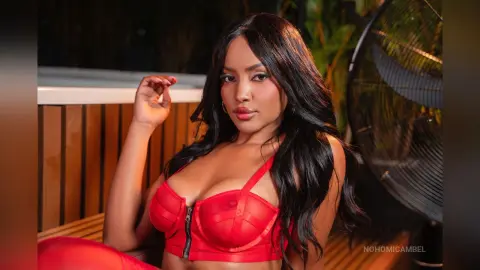 NohomiCambel poses in a red bra with black zipper detail, looking at the camera with long dark hair.