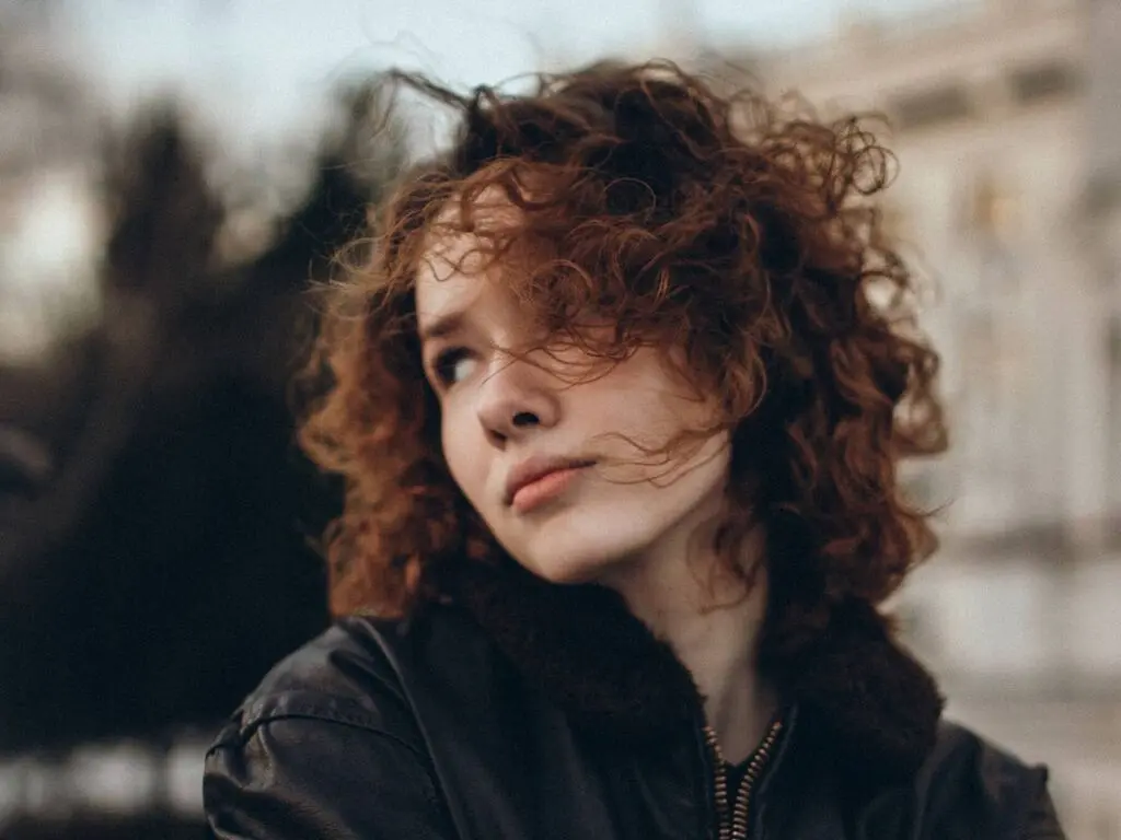 Young woman with curly red hair wearing a black jacket in a soft, moody outdoor setting.
