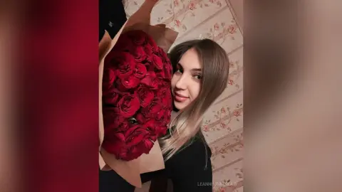Smiling young woman in a black top holding a massive bouquet of red roses against floral wallpaper.