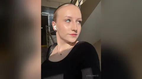 Young woman with slicked-back hair wearing a black top and wireless earbuds in soft sunlight.