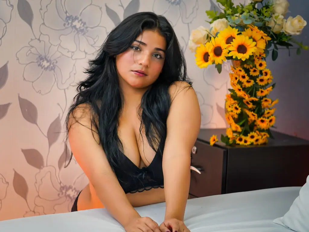 Young woman in black lingerie sitting by sunflowers against floral wallpaper in a bright, cozy room.