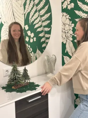 Smiling woman in a beige sweatshirt looking in a mirror against botanical green wallpaper in a bright room.