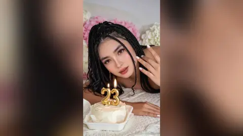 Young woman with long braids celebrating her 23rd birthday with a white cake in a soft, dreamy setting.