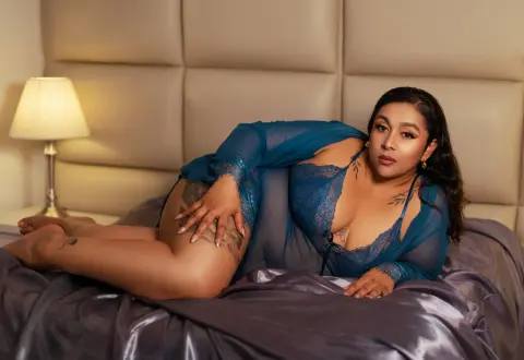 Curvy woman in blue lace lingerie relaxing on a bed in a warm, moody bedroom setting.