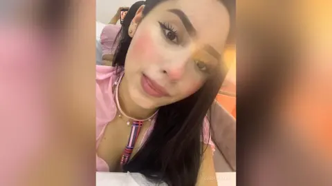 Young woman with long dark hair wearing a pink top and colorful necklace in a soft indoor setting.