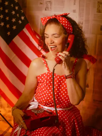 Smiling woman in a red polka dot dress using a vintage red telephone against an American flag background.