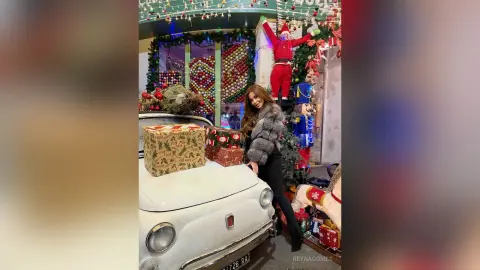 ReynaGomes poses next to a white car decorated with Christmas gifts and holiday lights, wearing a grey fur jacket and black pants.