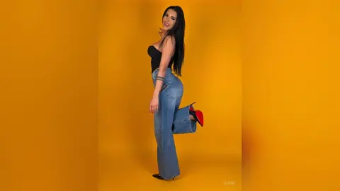 Smiling woman in black crop top and blue jeans posing against a vibrant yellow background.
