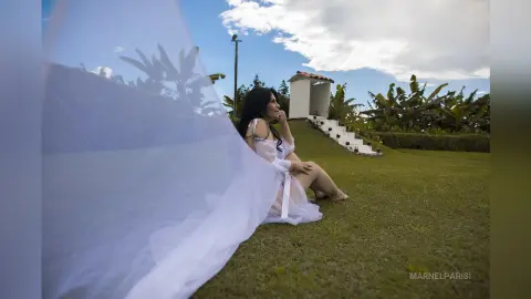 Dreamy portrait of a woman in a white flowing dress sitting on a grassy hill under a bright blue sky.