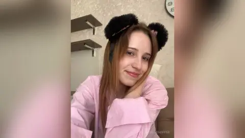 Smiling young woman in a pink shirt wearing fuzzy black cat ear headbands in a cozy indoor setting.