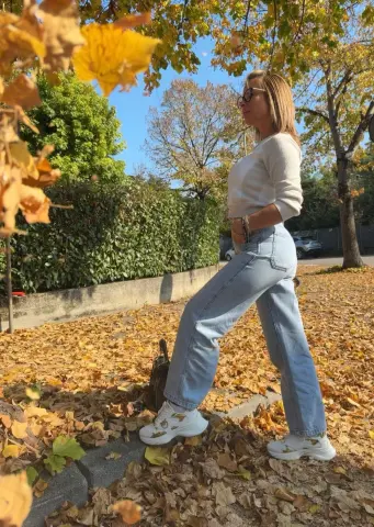 Woman in white sweater and light blue jeans walking through autumn leaves in a sunny park.