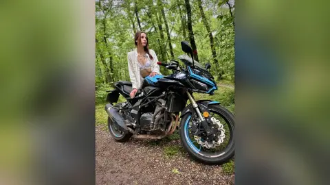 Stylish woman in a white jacket sitting on a black sport motorcycle in a lush green forest setting.