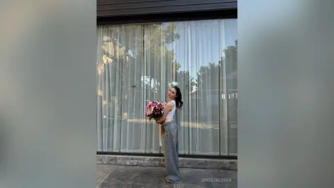 Young woman in a white tank top and baggy jeans holding pink flowers in front of a modern glass building.