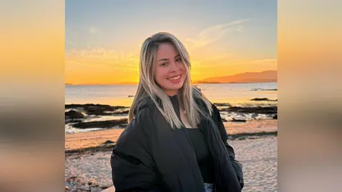 Smiling young woman in a black puffer jacket enjoying a golden sunset at the beach.