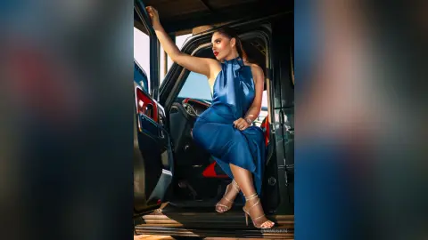 Elegant woman in a navy blue satin dress posing confidently in an open black vehicle during golden hour.