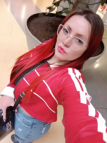 Red-haired woman in a red Adidas track jacket and jeans posing indoors with a casual, confident mood.