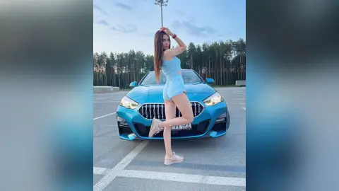 MilanaVolkova poses in front of a blue car, wearing a light blue crop top and matching shorts with her hands in her long brown hair.