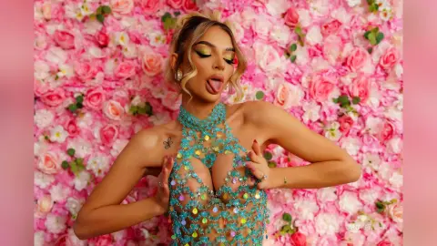 Anastasia poses against a pink rose wall wearing a sheer, turquoise sequined halter top that reveals her nipples.