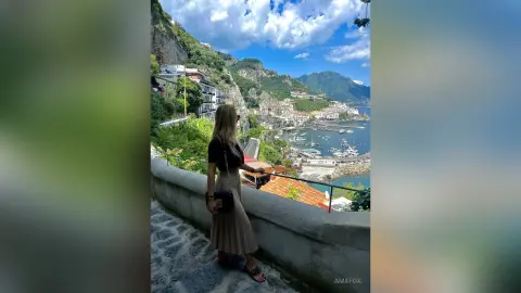 AmaFox — Blonde woman in a black top and pleated skirt overlooking the scenic Amalfi Coast seaside town.