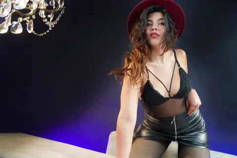 Glamorous woman in black lace lingerie and leather skirt posing under a crystal chandelier in moody lighting.