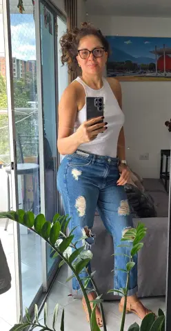 Smiling woman in a white ribbed tank top and distressed blue jeans posing in a bright sunlit room.