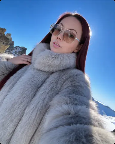 Young woman in a cozy grey faux fur coat posing against a bright blue winter sky in the snowy mountains.