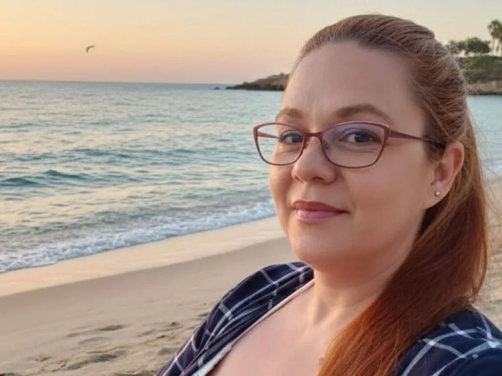 VictoriaDeea — VictoriaDeea smiles at the camera with her red hair pulled back, wearing glasses and a blue and white plaid shirt on a beach at sunset.
