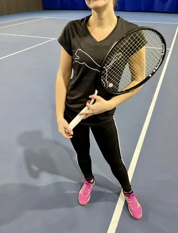 misssLush — misssLush stands on a blue tennis court wearing a black v-neck t-shirt, black leggings, and pink sneakers while holding a tennis racket.