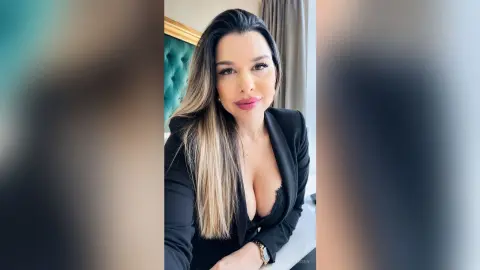 CapriAndersen wearing a black blazer with a low-cut neckline, showing her cleavage and long blonde-highlighted hair.