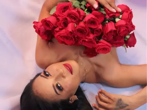 Luisa_Milf lies on white sheets, holding a large bouquet of red roses against her bare chest, showing her face and dark hair.