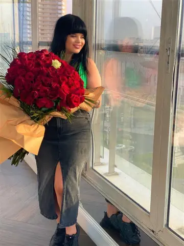 Smiling woman in a green top and denim skirt holding a large bouquet of red roses by a city window.