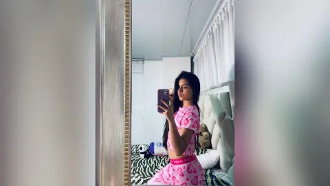 Young woman in pink patterned loungewear taking a mirror selfie in a bright, modern bedroom.