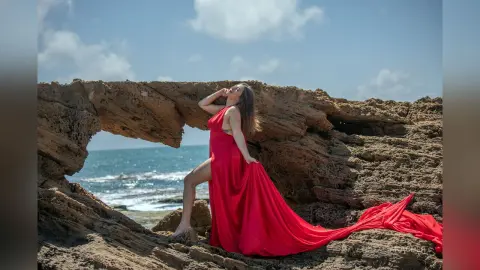 NataliaNacher poses in a long red dress against a rocky coastline with the ocean and blue sky in the background.