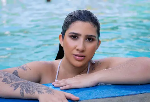 Tattooed woman in a white bikini relaxing by a turquoise swimming pool on a sunny summer day.