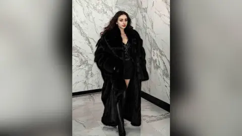Elegant woman in a luxurious black faux fur coat and black dress posing against a marble wall.