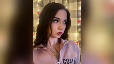 Young woman with long wavy hair wearing a pink zip-up sweatshirt in a cozy room with fairy lights.