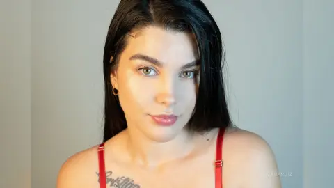 Young woman with long dark hair and glowing skin wearing a red strap top in a minimalist indoor setting.