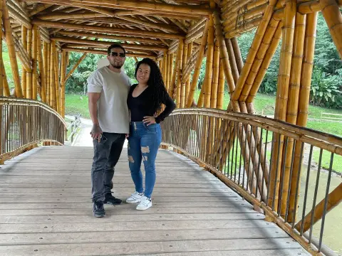 Smiling couple in casual outfits standing on a rustic bamboo bridge in a lush tropical garden setting.