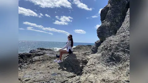 Woman in white shirt and shorts sitting on rocky cliffs overlooking the blue ocean under a sunny sky.