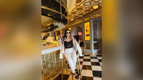 Stylish woman in white trousers and black lace bodysuit posing in a luxurious gold and black patterned lounge.